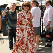 Person in floral maxi dress with sunglasses walks outdoors, holding a phone. Others casually stroll nearby, including a person giving a thumbs-up