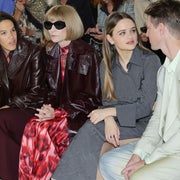 Four people seated at a fashion show. One in a long leather coat, another in a long dress, next in a blazer, and the fourth in a suit