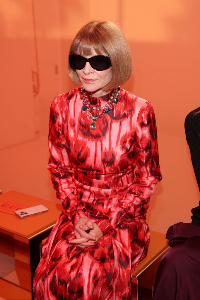 Person in a patterned dress with sunglasses sits at an event, wearing a statement necklace