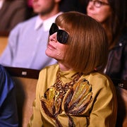 Person seated at an event, wearing sunglasses and a gold top with leaf embellishments, observing intently