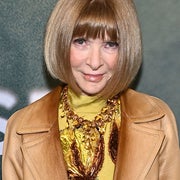 Person with short bob hairstyle wearing a leather jacket over a detailed, high-neck top adorned with a large, ornate necklace