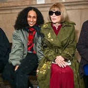 Two people seated; left person in casual wear, right person in a floral trench coat and sunglasses, often seen at fashion events