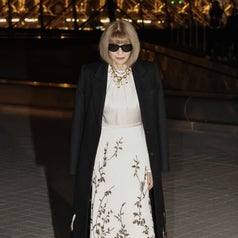 Person in a floor-length floral gown with a black overcoat and sunglasses, standing outdoors under an illuminated structure