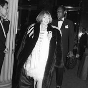 Person in a fur coat over a short dress, attending an event with elegantly dressed people nearby
