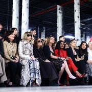 Fashion show audience watches a model walking the runway in a sequined full-length outfit with a high collar. Attendees wear stylish, diverse outfits