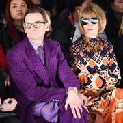 Individuals seated at a fashion event. One wears a purple striped suit, and another dons a patterned dress with large sunglasses