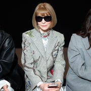 Person with sunglasses wearing a patterned blazer and floral dress sits in a row, holding a phone