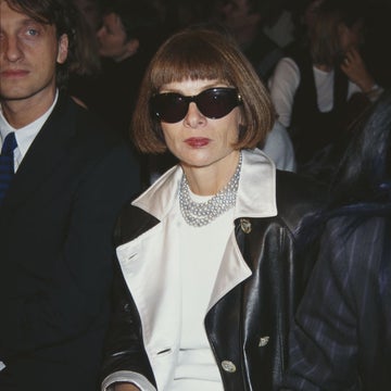 A person sits at a fashion event, wearing sunglasses, a white outfit, and a black leather jacket with a pearl necklace