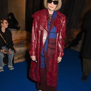 Person wearing large sunglasses, a patterned red coat, a blue scarf, a red dress underneath, and snakeskin boots at a fashion event
