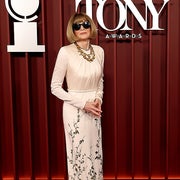 An elegant woman in a floral, floor-length gown, wearing sunglasses, stands on the red carpet at the Tony Awards
