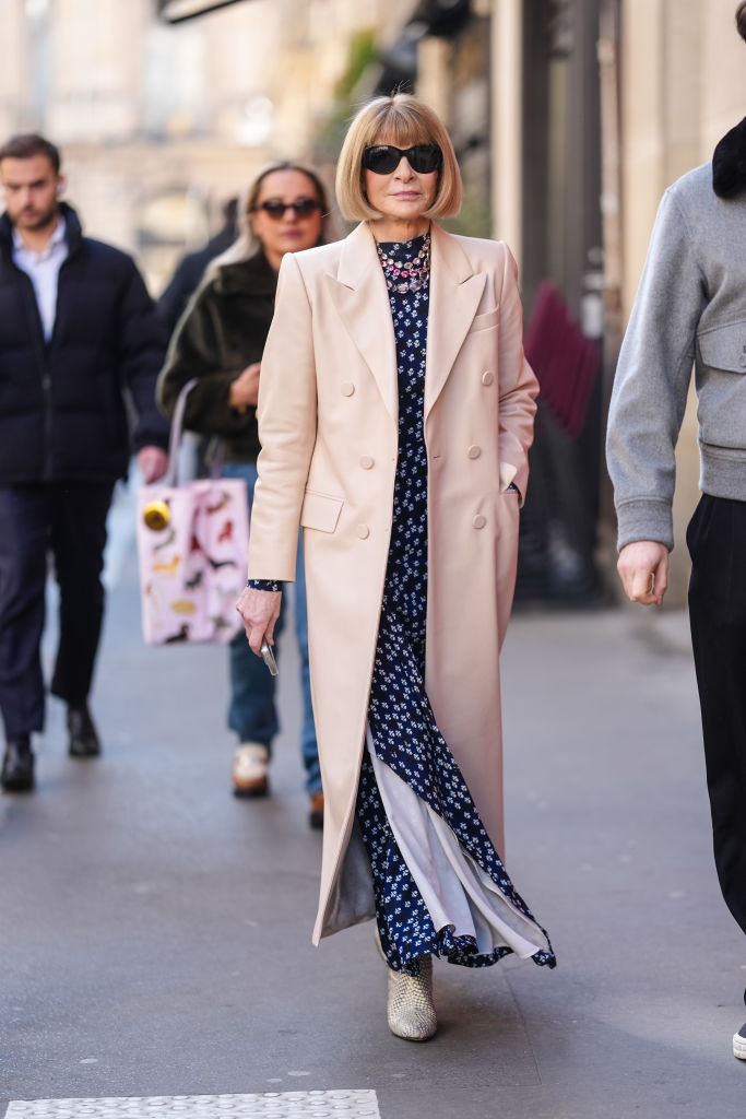 Person in a chic floral dress and long overcoat walks down a city street, flanked by people in casual wear
