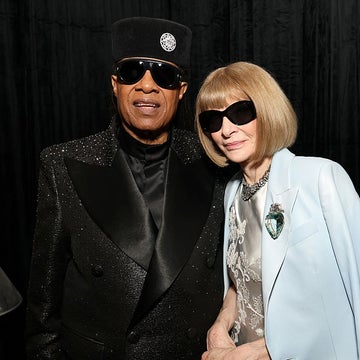 Two people wearing sunglasses and stylish outfits stand together at an event, one in a sparkling suit and hat, the other in a light-colored coat