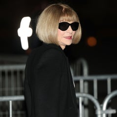 Person with bob haircut and sunglasses wearing a sleek black blazer, standing near a metal railing