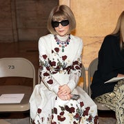 Person in a floral patterned dress, large sunglasses, and a statement necklace sitting on a chair indoors