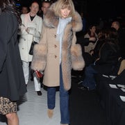 Person in a fur-trimmed coat walks on a white runway, surrounded by people in formal clothing