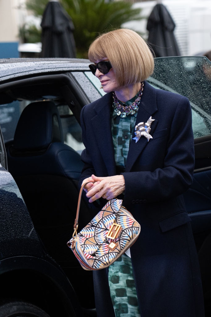 Person in dark coat with statement brooch and sunglasses holds a patterned handbag next to a car