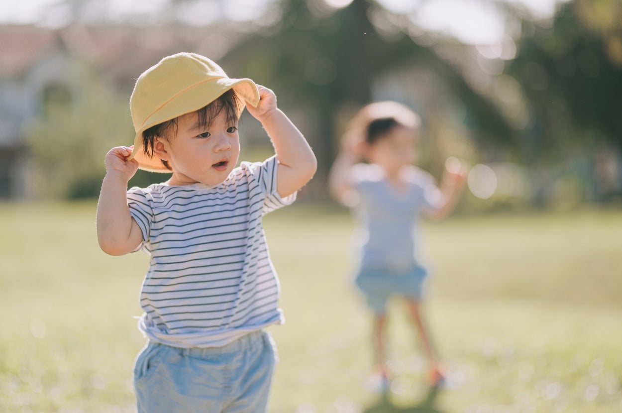 帽子をかぶった小さな子供が、芝生の上で遊ぶ様子。背景にはぼかされたもう一人の子供が見える。