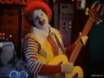 Character wearing clown makeup and costume, playing a large guitar animatedly on a colorful stage