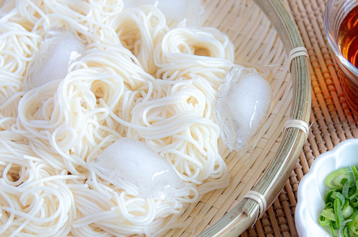 ザルに盛られた冷たいそうめん、氷が載せられている。横にめんつゆと薬味のねぎと生姜が添えられている。