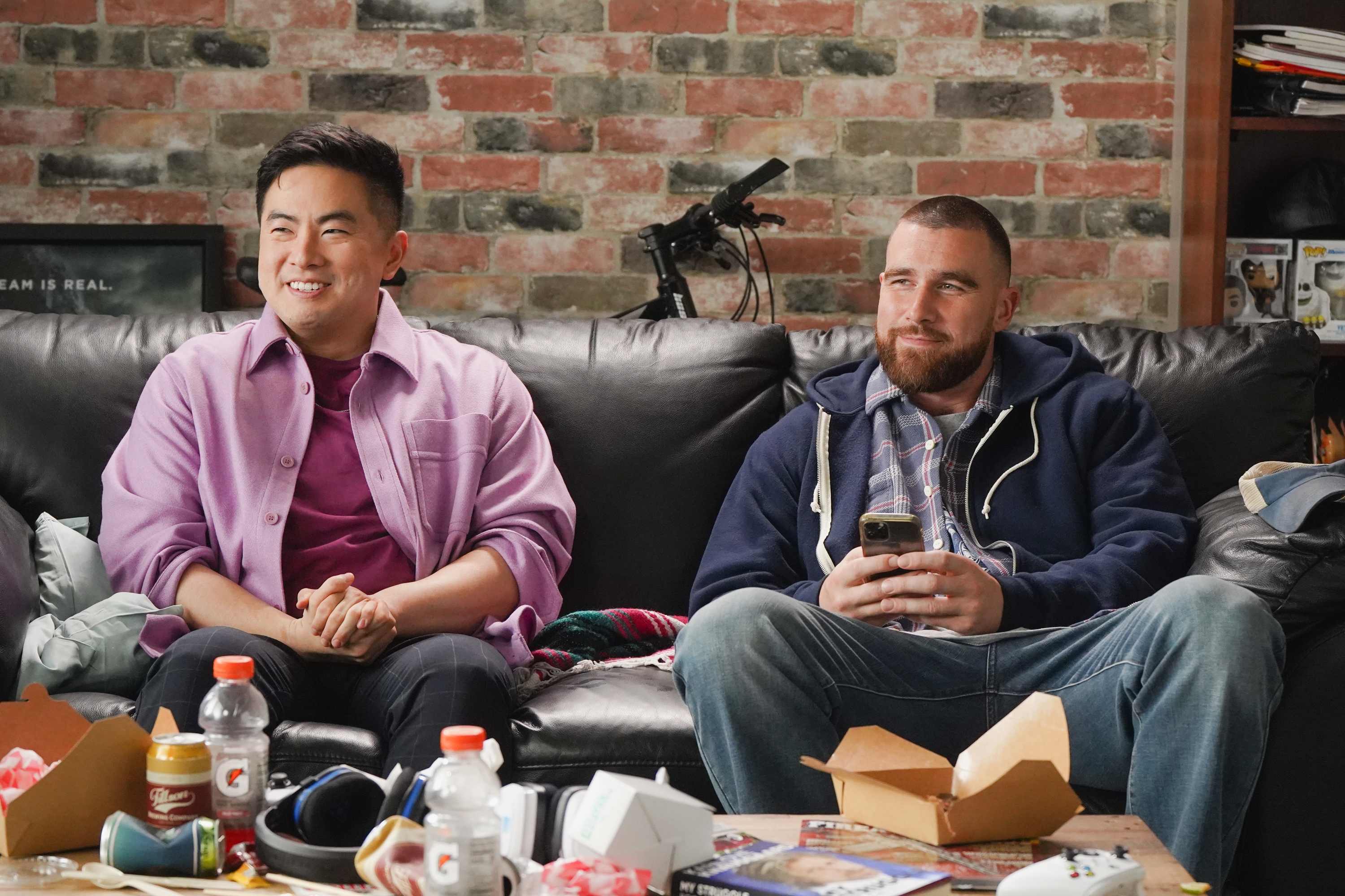 Two people sitting on a couch, one holding a phone. Casual attire, takeout food and drinks on the table. Cozy, relaxed setting