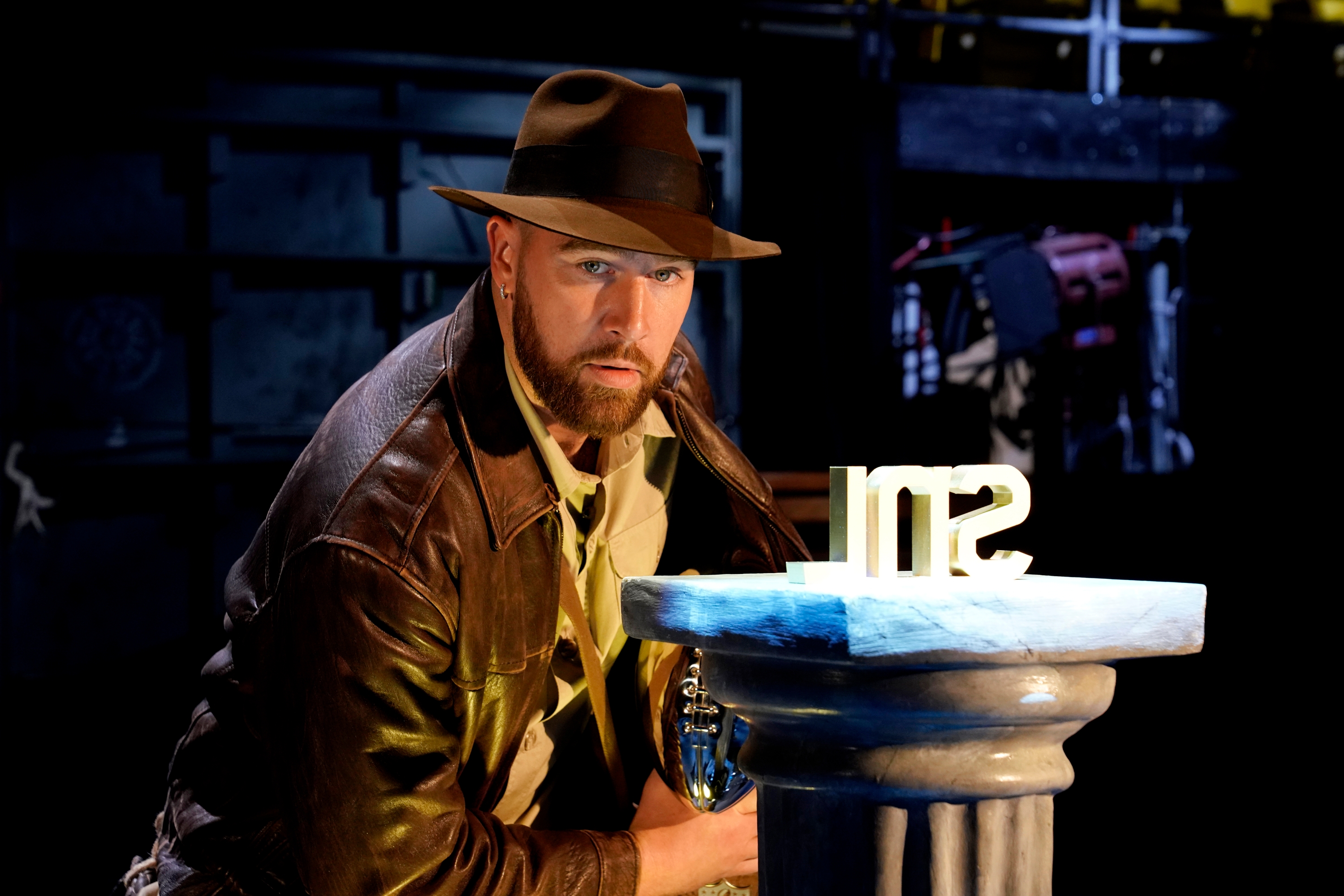 Man in an adventurer outfit with a fedora and leather jacket posing dramatically beside a pedestal with a white object