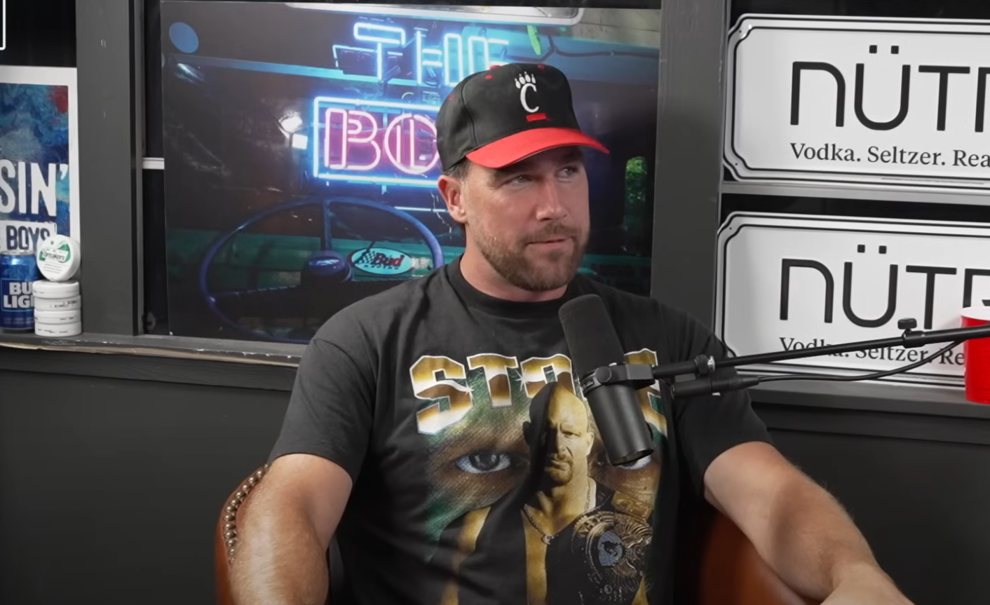 Person wearing a black cap and graphic T-shirt, sitting during a podcast interview with microphones visible. Neon sign in background