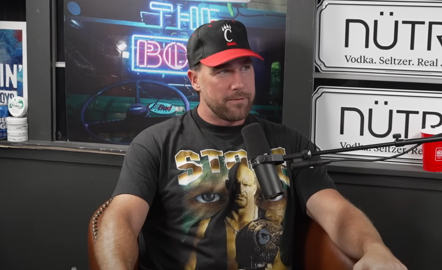 Person wearing a black cap and graphic T-shirt, sitting during a podcast interview with microphones visible. Neon sign in background