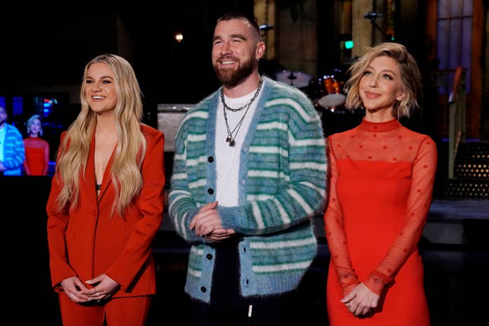 Three people smiling on a TV set; woman in red tailored suit, man in a striped cardigan, and another woman in a red mesh dress
