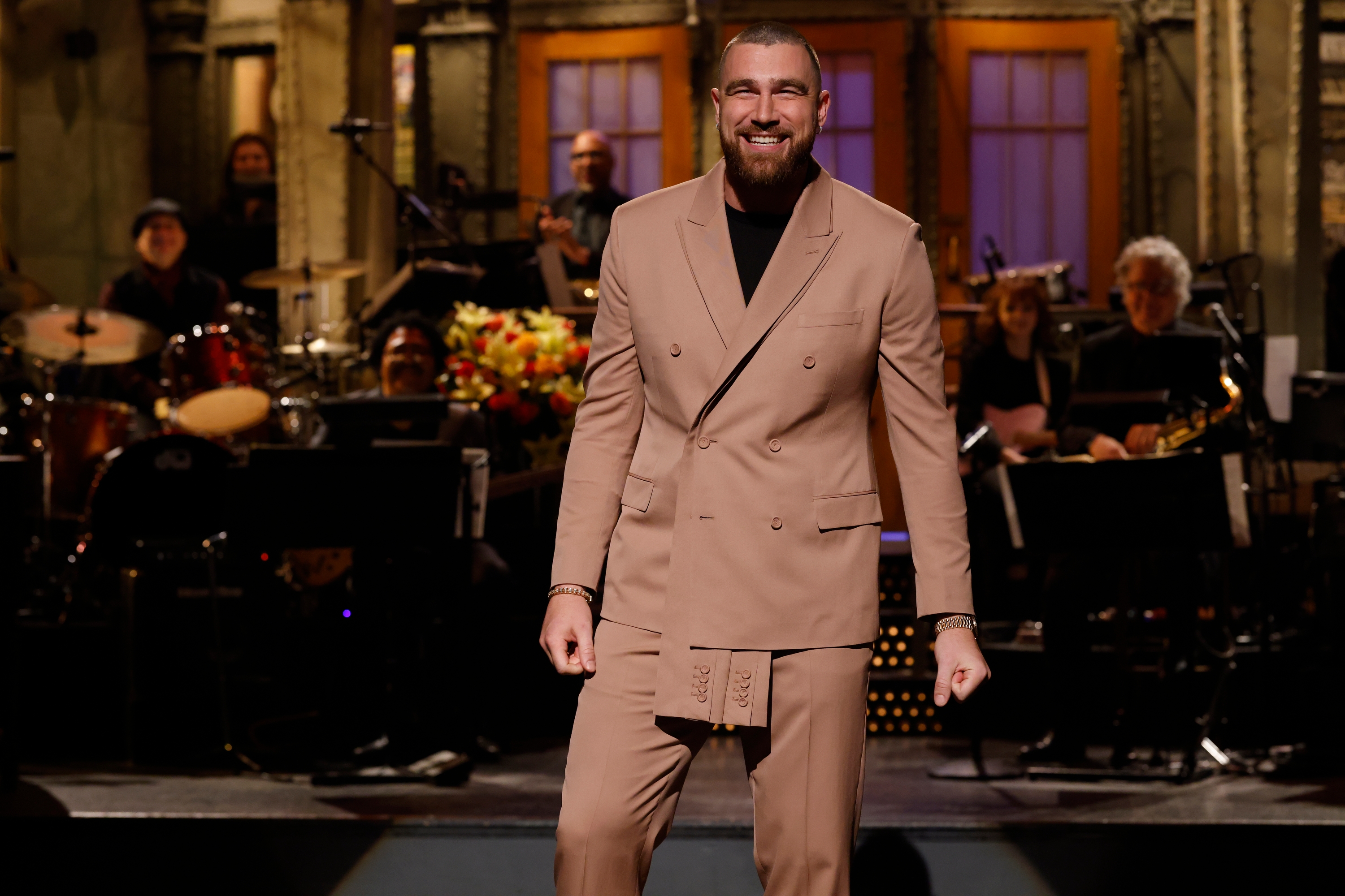 Person in a double-breasted suit smiles on stage with a band in the background during a live TV show
