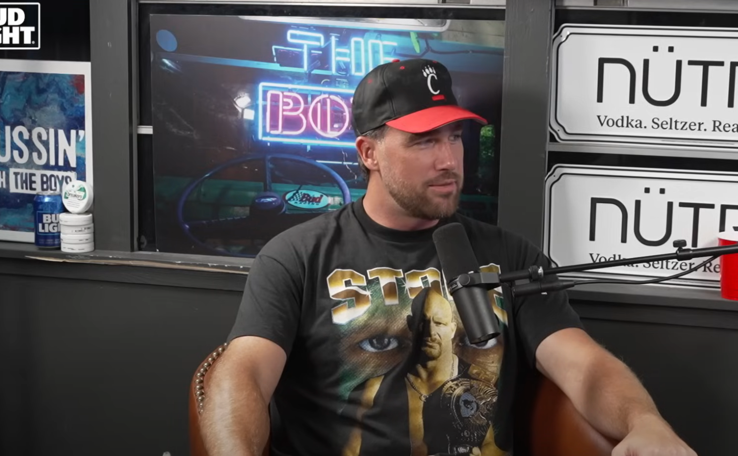 A person in a cap and graphic t-shirt sits during a podcast recording, microphone in front, with brand logos visible in the background