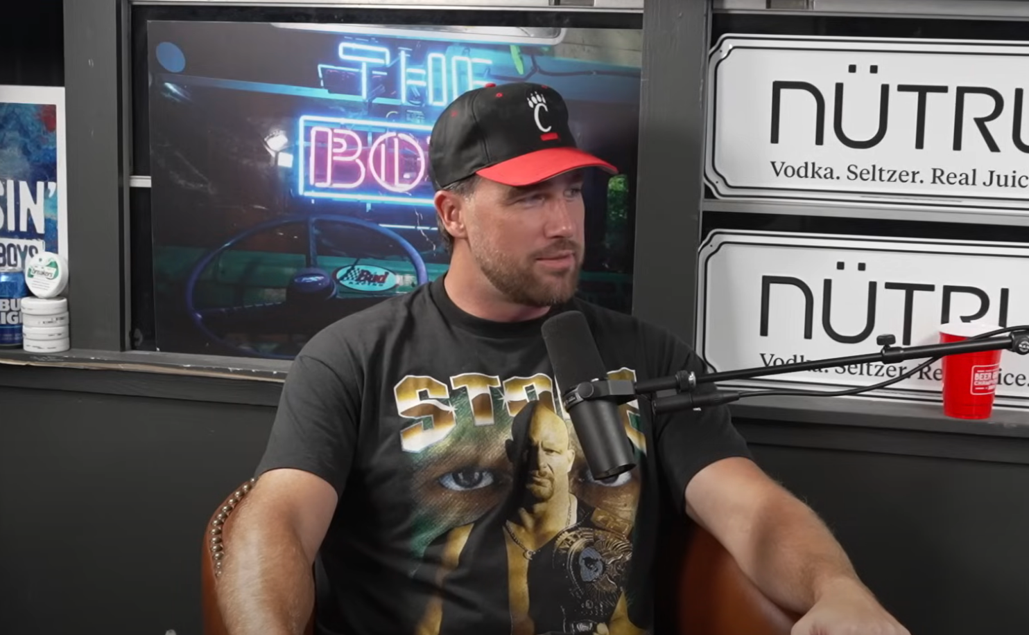 A person in a cap and graphic t-shirt sits during a podcast recording, microphone in front, with brand logos visible in the background