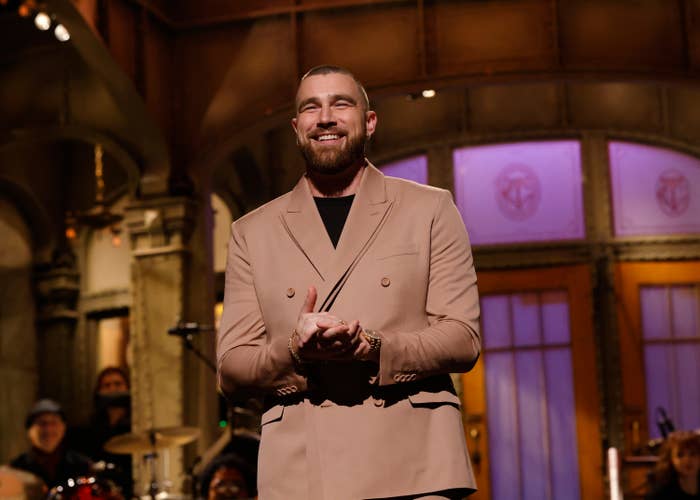 A man stands smiling on a stage, wearing a double-breasted suit jacket. The setting suggests a live performance or talk show