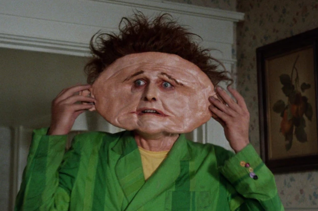 Person in a green outfit holds a large, exaggerated ear mask over their head in a humorous scene