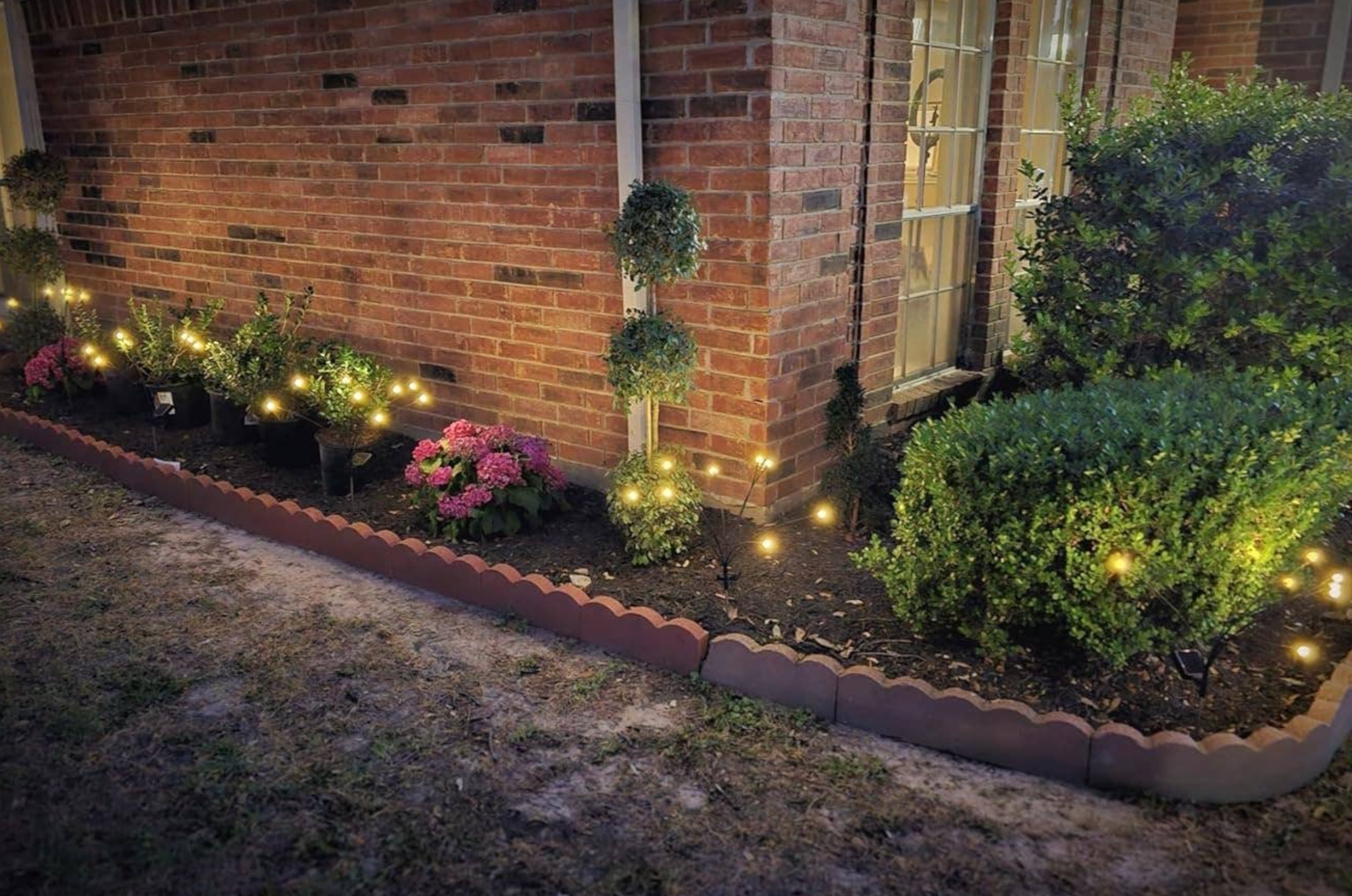String lights illuminate a garden bed with shrubs and pink flowers against a brick house, creating a cozy outdoor ambiance