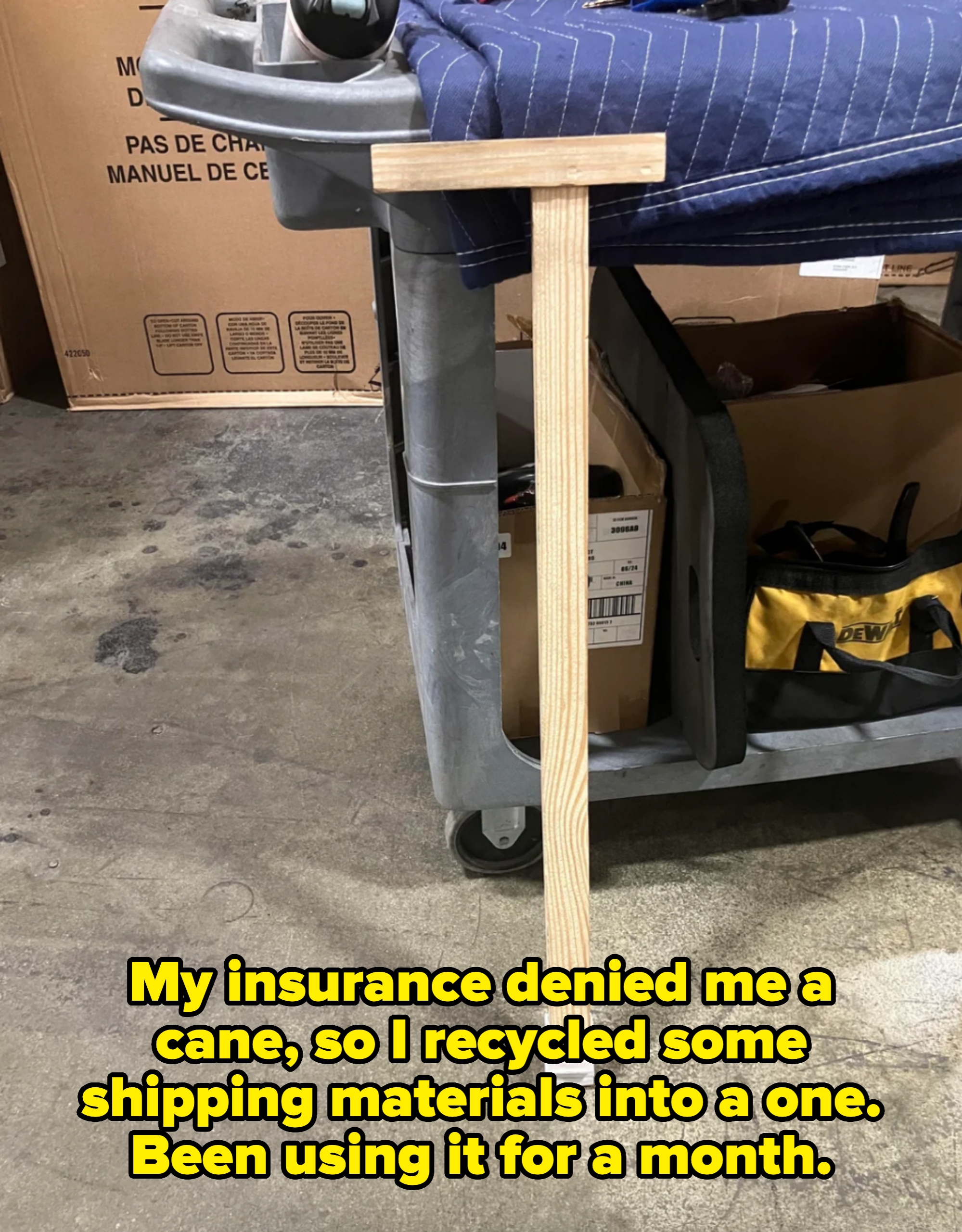 Wooden tool with a T-shape handle leaning against a cart in a warehouse setting. Caption: &quot;My insurance denied me a cane, so I recycled some shipping materials into a one. Been using it for a month&quot;