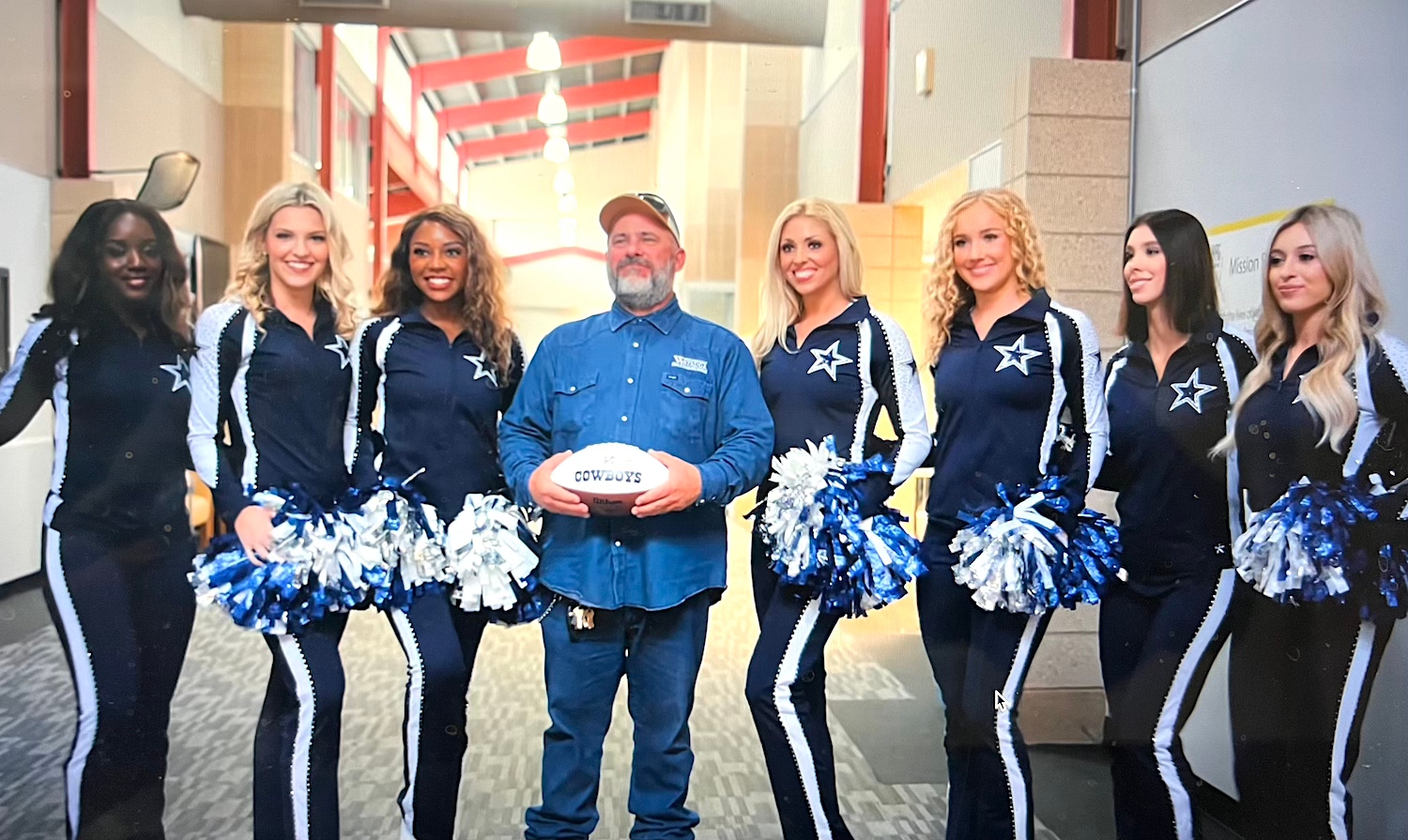 A person stands holding a football, surrounded by cheerleaders in matching outfits with star emblems, posing for a photo