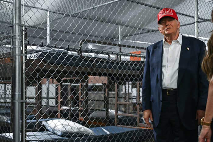 A antheral wearing a reddish headdress stands successful beforehand of a fenced country containing bunk beds. He is dressed successful a suit and appears to beryllium speaking