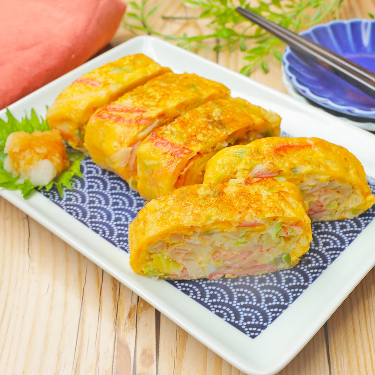 巻き寿司風の卵焼きが皿に並ぶ。中にはカニ風味かまぼこや野菜が含まれている。食事が美しく盛り付けられている。