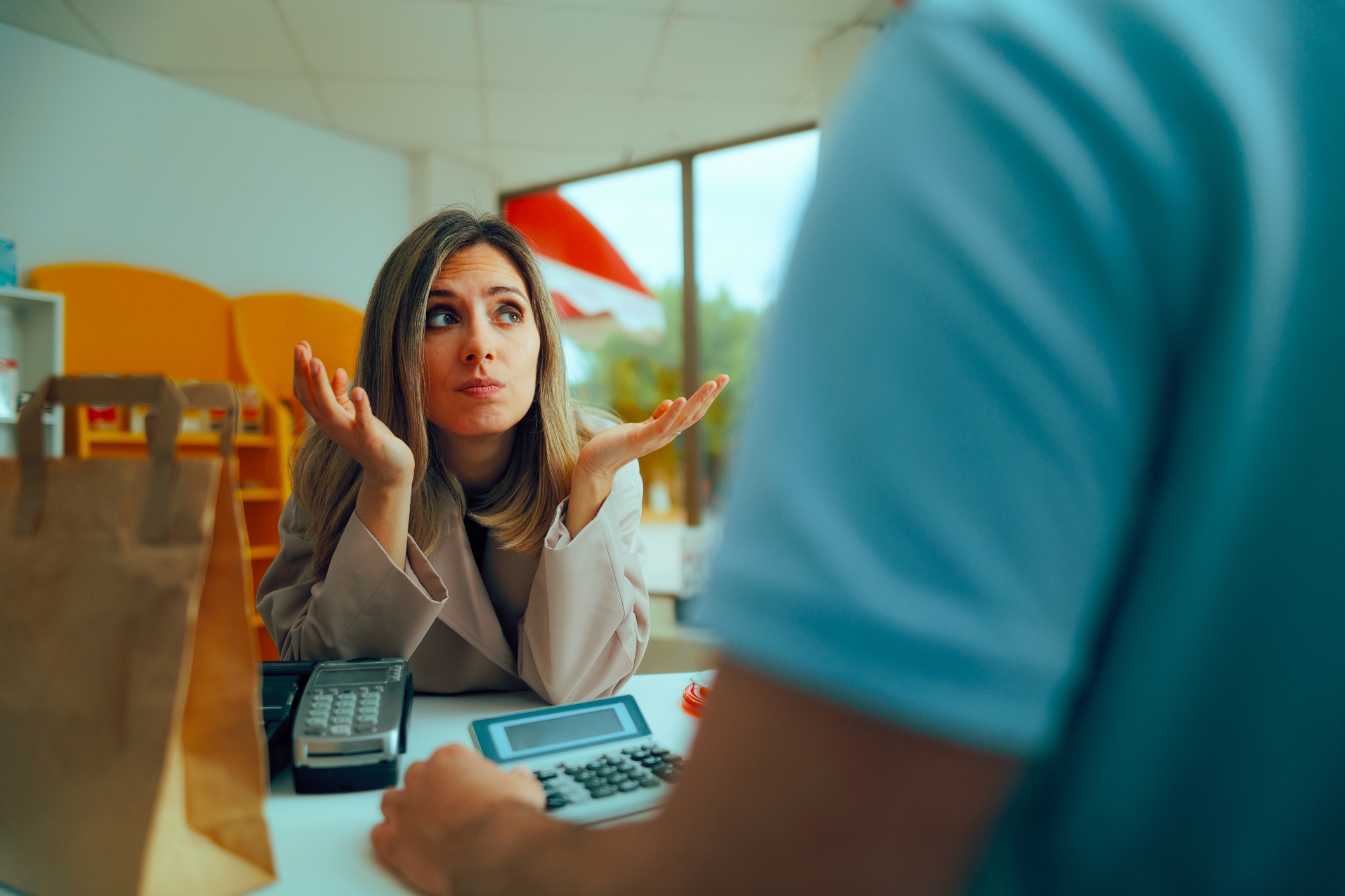 Kvinne ved en teller, trekker på skuldrene med håndflatene, ser forvirret ut. Brun papirpose og kalkulator synlig på disken