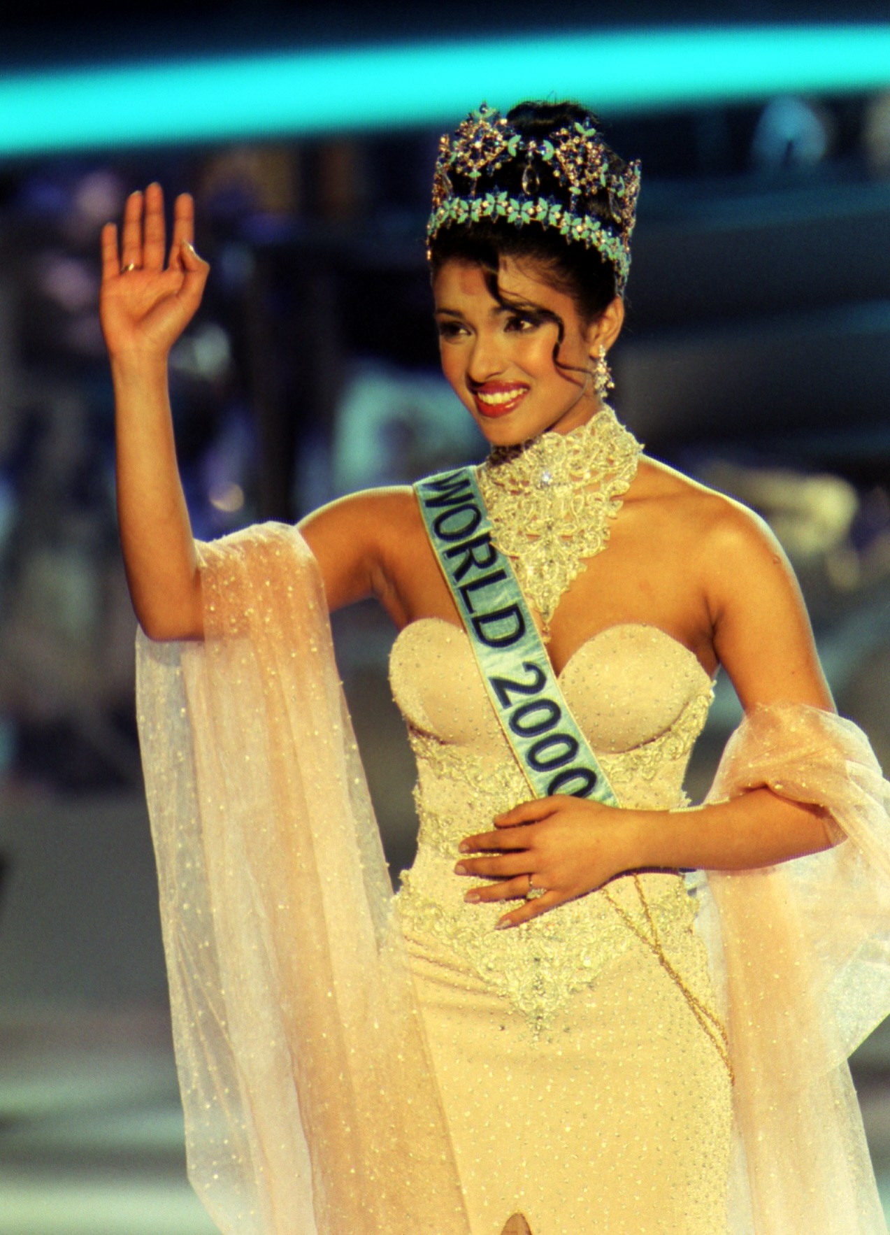 Person wearing an ornate gown and a crown, with a sash reading "World 2000," waves to the audience