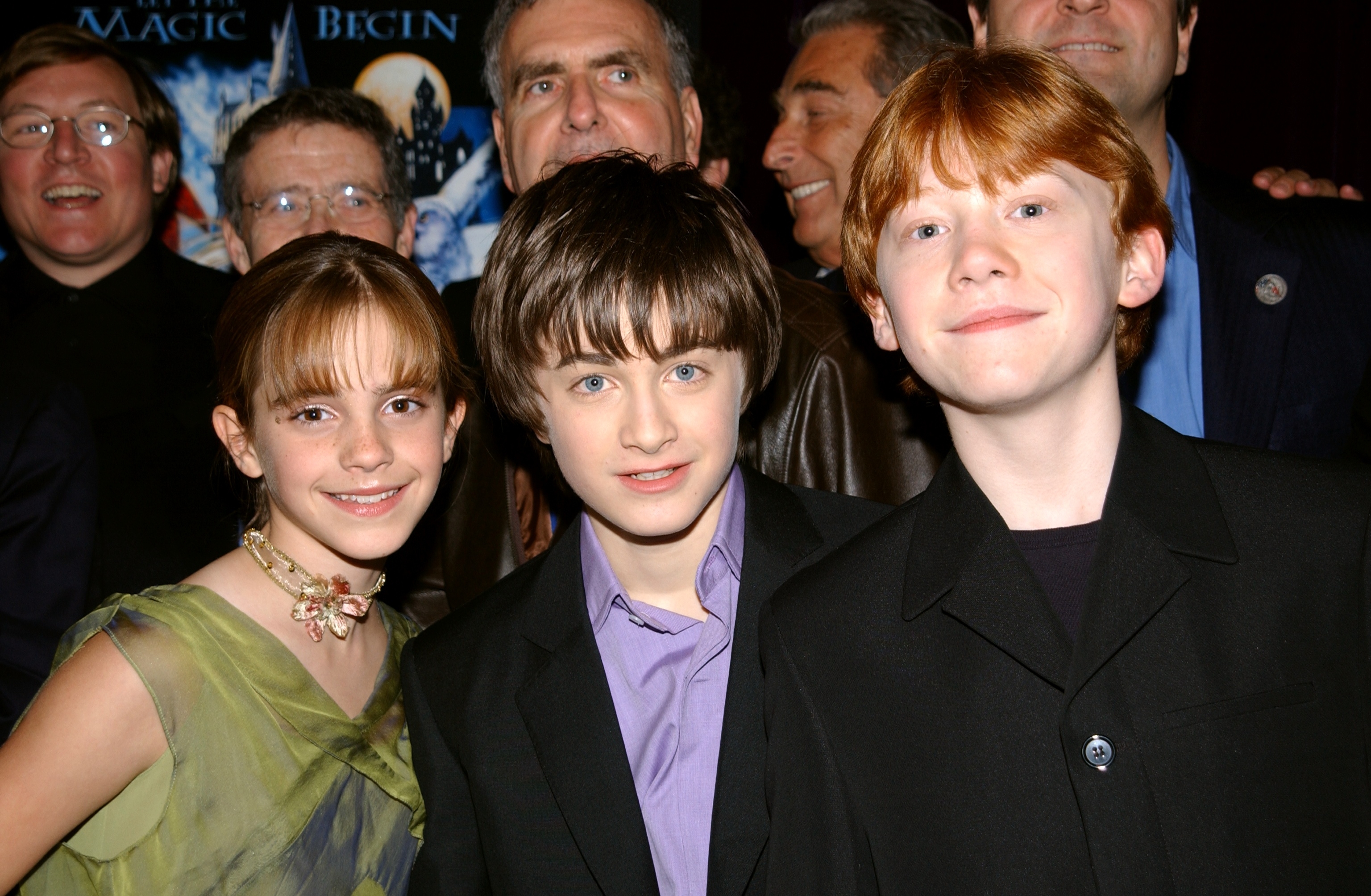 Three young actors in formal attire pose at a movie event with a fantasy theme backdrop and smiling people in the background