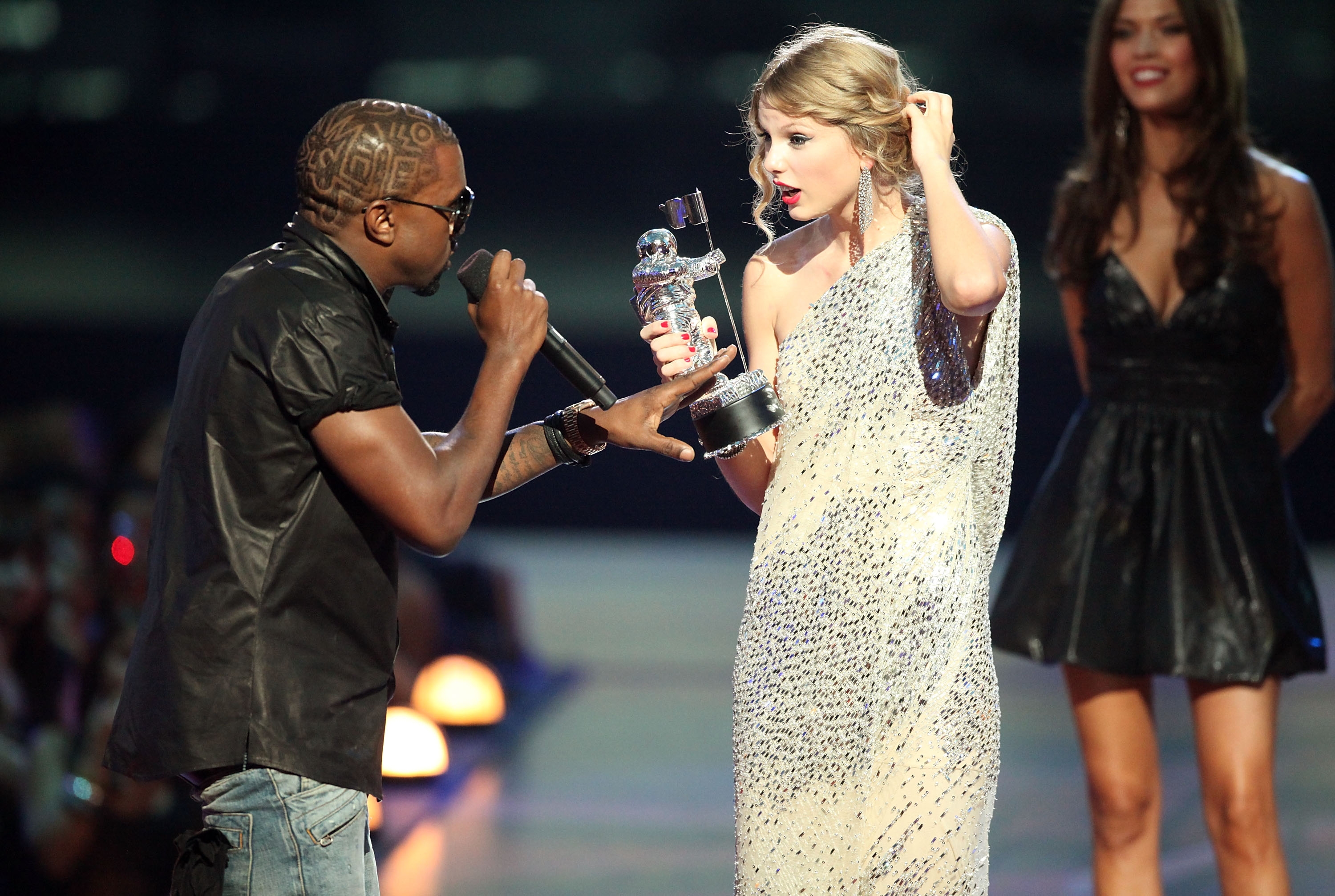 Taylor Swift holds an award in a sparkly dress, surprised, as a man gestures and talks on stage. Another woman in a short dress stands nearby