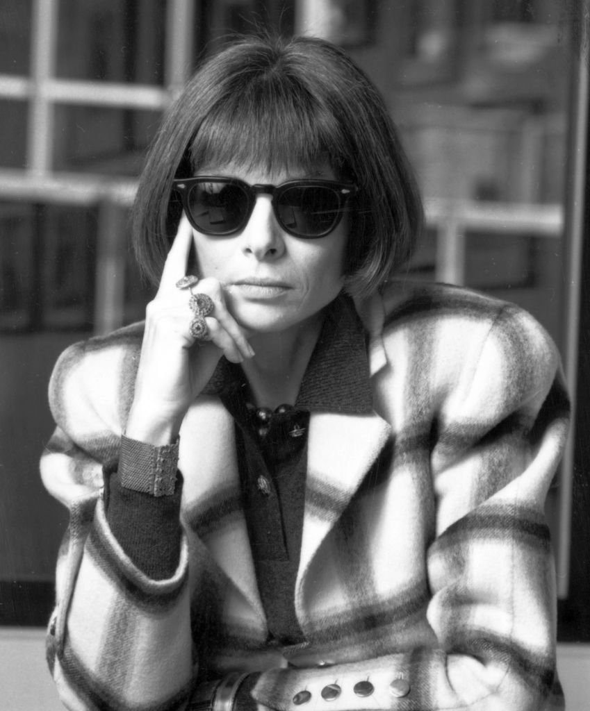 Person wearing a checkered jacket, sunglasses, and resting chin on hand in a thoughtful pose