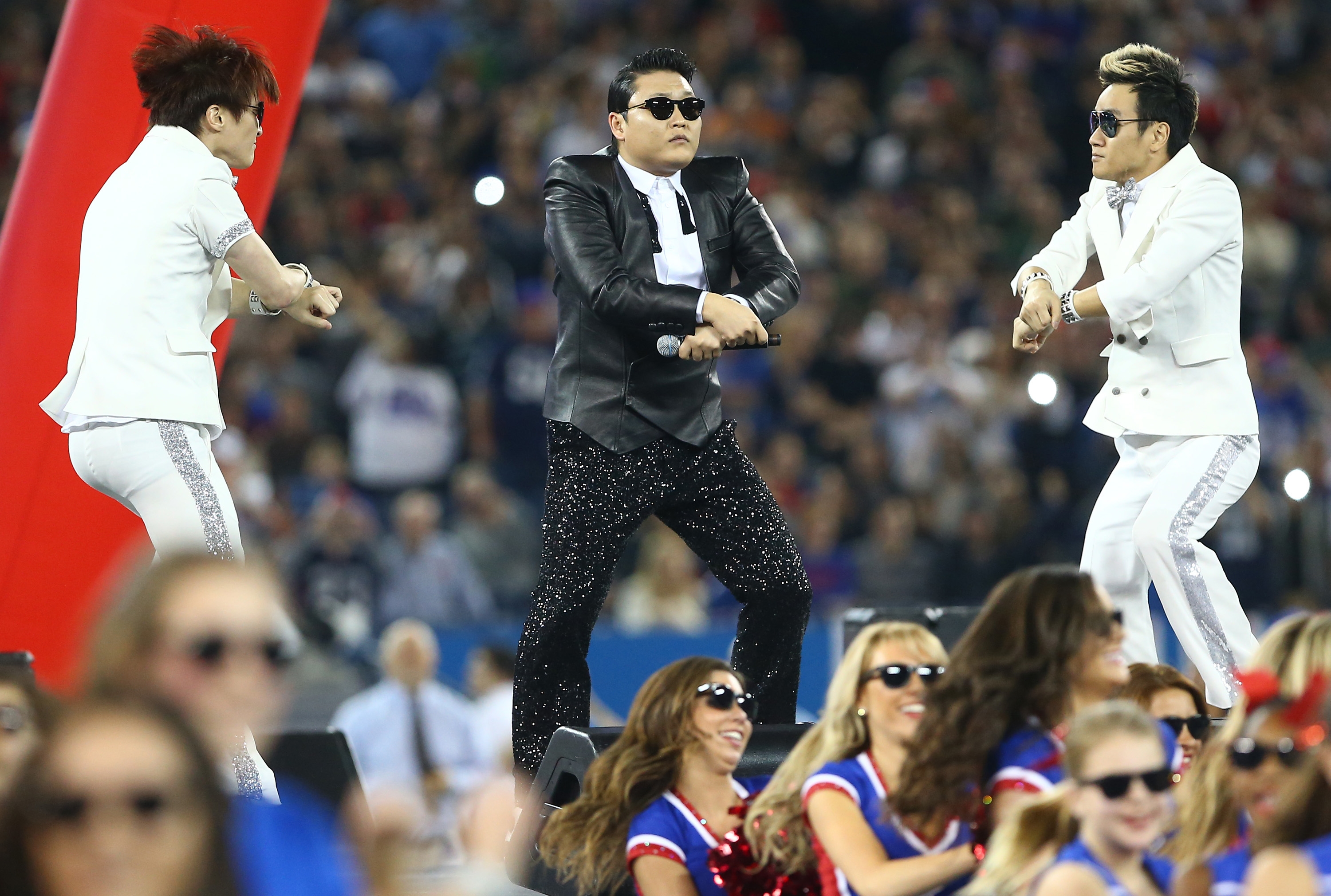 Performer in a black sequin outfit dances on stage with male dancers in white suits, surrounded by cheerleaders