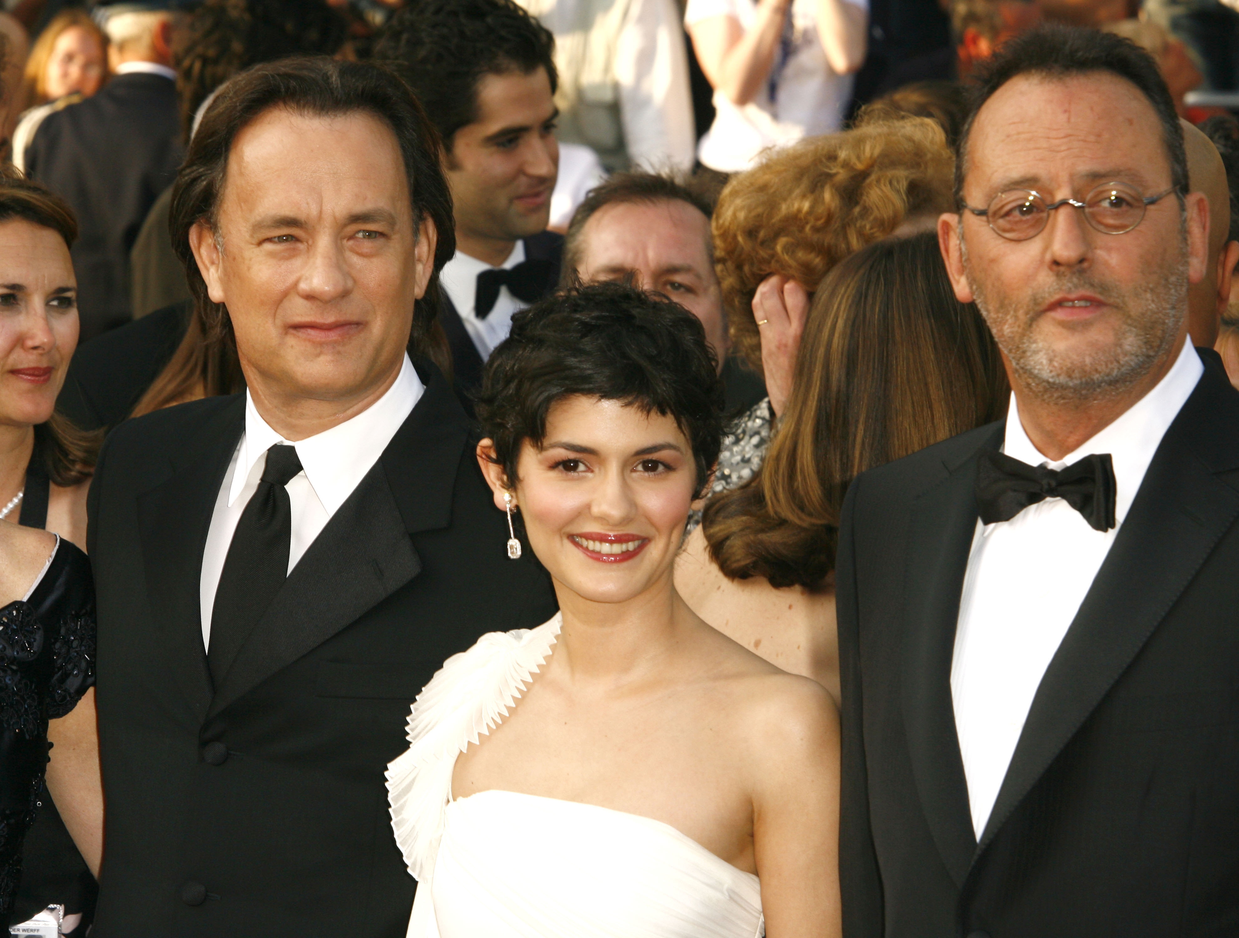 Three people on a red carpet in formal wear with suits and an off-shoulder dress, posing for a photo