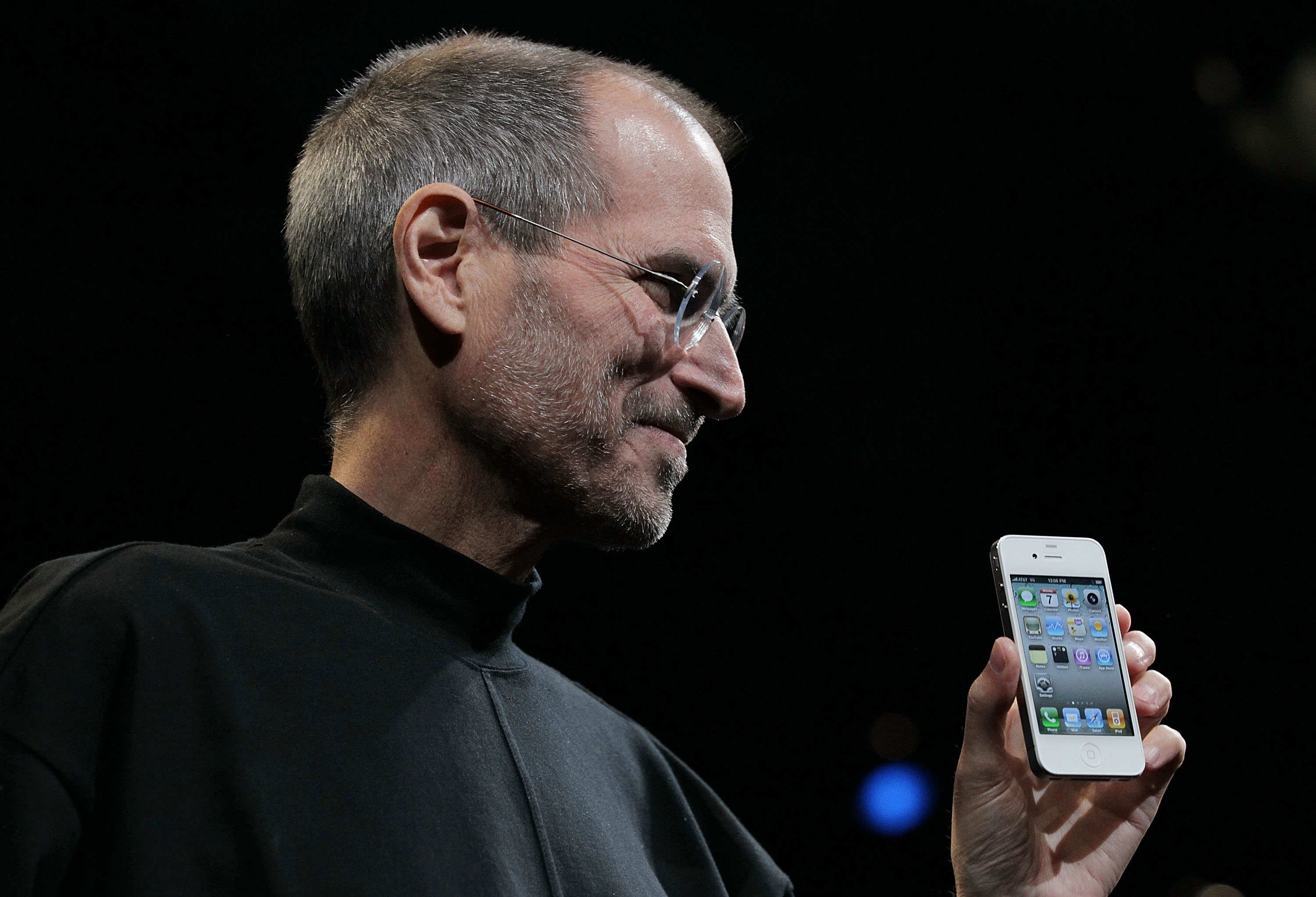 A person holding a smartphone on stage, wearing a simple black turtleneck and glasses