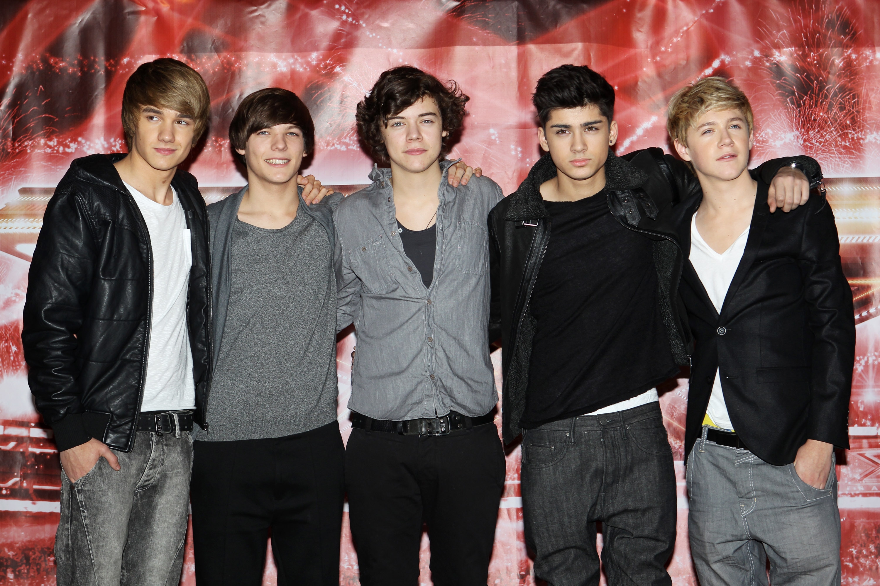 Five young men pose together, wearing casual outfits with jackets, tee shirts, and jeans, in front of a red carpet backdrop
