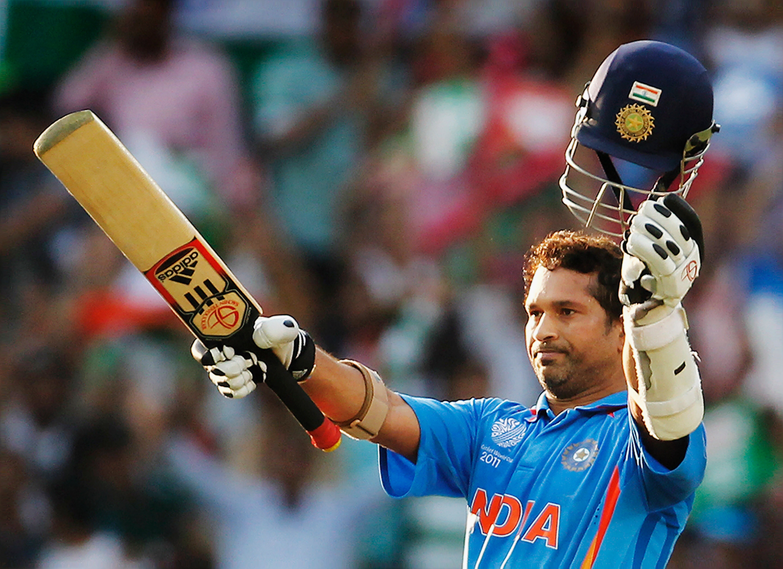 Cricketer in blue uniform raises a bat and helmet in celebration on the field