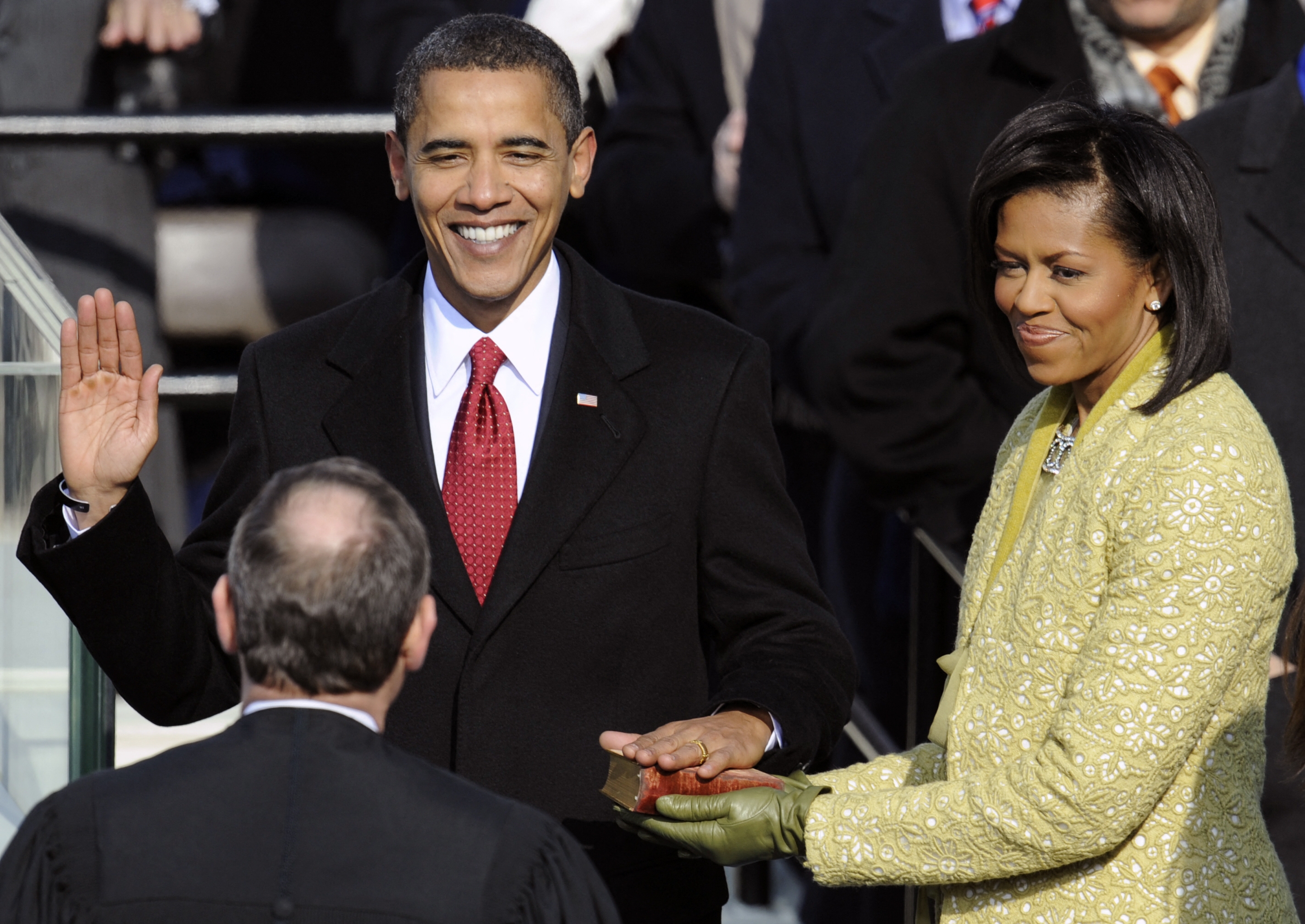 A person is being sworn in, wearing a suit with a tie; another stands beside, holding a Bible. They are on an official platform with a judge present