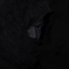 A person peeks out from under a rough, dark hood, partially concealing their face in shadows